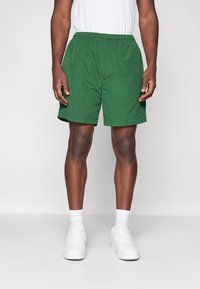 Pantalones cortos deportivos verdes hechos de una tela ligera; cuentan con una cintura elástica y bolsillos laterales; el dobladillo llega por encima de la rodilla.