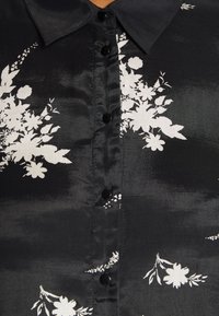 Camicia di tessuto nero con motivo floreale bianco, dotata di colletto a punta e bottoni neri lungo la parte anteriore. Texture liscia.