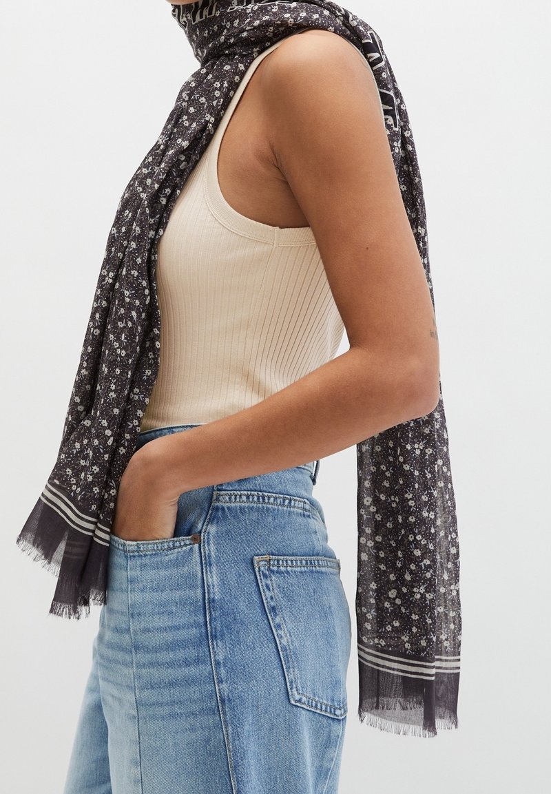 Personne portant un haut sans manches côtelé crème, un jean taille haute bleu avec une main dans la poche, et une écharpe à motif floral sombre autour du cou.
