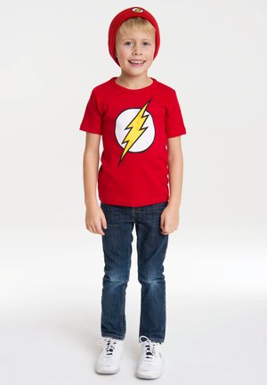 Rotes T-Shirt mit kurzen Ärmeln und einem gelben Blitzlogo auf einem weißen Kreis, kombiniert mit blauen Jeans und weißen Turnschuhen. Rote Mütze inklusive.