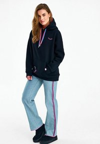 Pikkade juustega naine musta kapuutsiga, millel on värvilised nöörid, ja helesinised teksad roosa ja lilla küljeribaga, seisab ühe käega taskus.