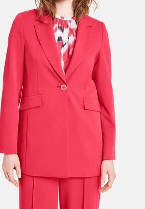 Blazer rosa con solapas con muesca, cierre de un solo botón, bolsillos frontales y un corte relajado. Usado sobre una blusa con estampado.