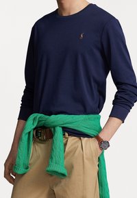 Mörkblå långärmad skjorta med rund halsringning, i kombination med en grön kabelstickad tröja knuten runt midjan och beige byxor. Brun armbandsklocka synlig.