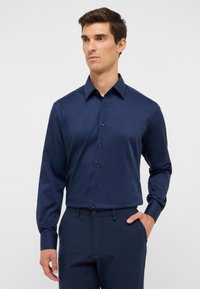 Uomo vestito con una camicia a maniche lunghe blu navy con bottoni e colletto, pantaloni blu navy, con una mano in tasca, su uno sfondo chiaro e semplice.