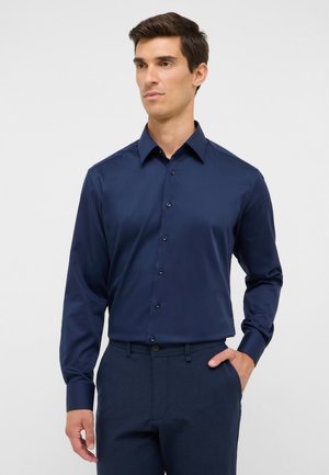 Mann trägt ein knöpfiges, dunkelblaues Langarmhemd mit Kragen und eine dunkelblaue Hose, eine Hand in der Tasche, vor einem einfarbigen hellen Hintergrund.