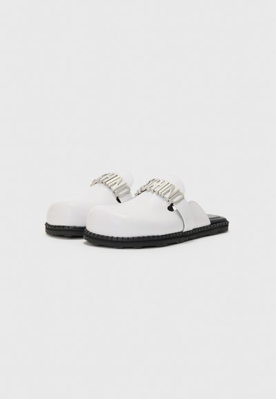Baltie slip-on apavi ar biezu, apaļu purngalu, ar melnu gumijas zolīti un sudraba logo akcentu uz siksniņas. Gluda apdare.