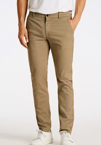 Pantalones caqui de tejido suave con corte slim. Cuentan con cremallera y bolsillos laterales, combinados con zapatillas blancas para crear contraste.