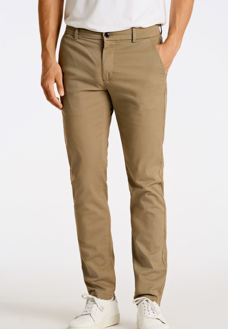 Pantalones caqui de tejido suave con corte slim. Cuentan con cremallera y bolsillos laterales, combinados con zapatillas blancas para crear contraste.