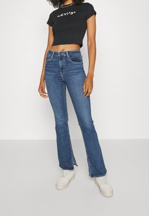 Woman wearing black cropped Levi's t-shirt, blue high-waist flared jeans with front slits, and white platform sneakers standing against white background.