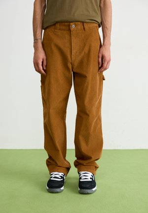 Pantalons cargo en velours côtelé marron avec une coupe droite, dotés de poches latérales, d'une fermeture à bouton, portés avec des baskets noires.