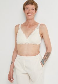 Brassière en dentelle ivoire avec des bords festonnés et un délicat nœud au centre. Le tissu présente des motifs floraux intricats.