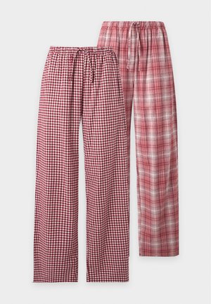 Femme aux longs cheveux foncés portant un haut blanc sans manches et un pantalon de pyjama large à carreaux rouges et blancs, debout pieds nus sur un fond blanc.