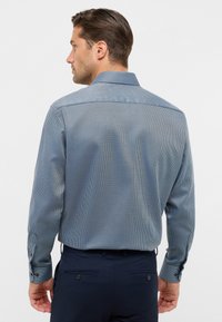 Uomo che indossa una camicia a maniche lunghe, blu con motivo, infilata in pantaloni blu scuri, visto da dietro su uno sfondo uniforme.