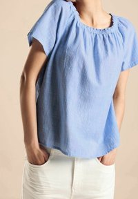 Jasnoniebieska bluzka z krótkim rękawem i zdobionym dekoltem, uszyta z teksturowanej tkaniny. W połączeniu z białymi spodniami, ręce włożone w kieszenie.