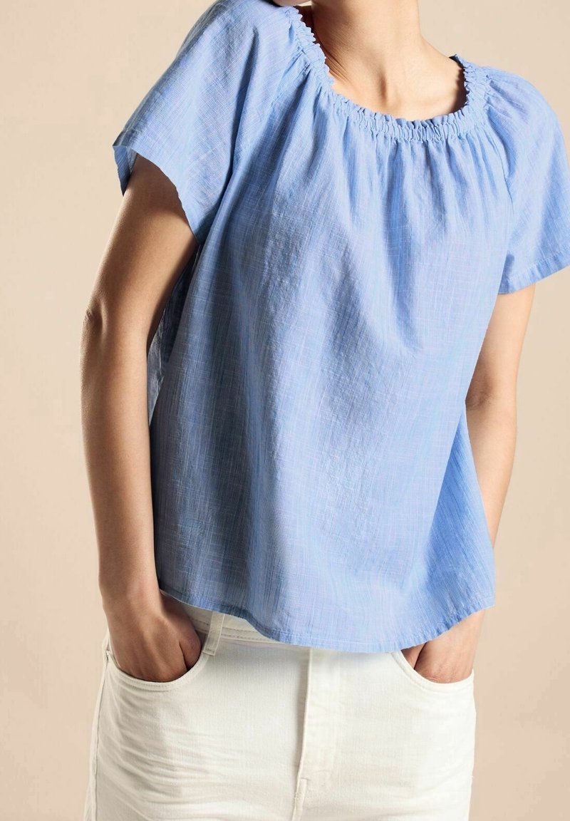 Jasnoniebieska bluzka z krótkim rękawem i zdobionym dekoltem, uszyta z teksturowanej tkaniny. W połączeniu z białymi spodniami, ręce włożone w kieszenie.