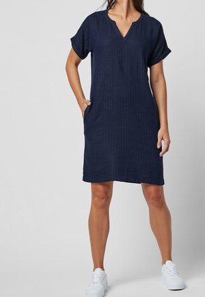 Femme portant une robe bleu marine à manches courtes, longueur genoux, avec des poches et des baskets blanches, debout devant un fond gris uni.