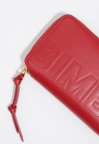 Czerwony skórzany portfel z wytłoczonym napisem. Posiada złoty zamek błyskawiczny oraz odpinany, węzłowy pasek do łatwego noszenia.