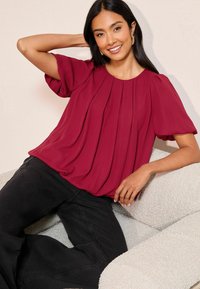 Burgundin värinen laskostettu paita lyhyillä puffihaitoilla, pehmeä tekstuuri, pyöreä kaula-aukko, yhdistetty mustiin leveneviin farkkuihin, asetettu teksturoidulle vaalealle pinnalle.