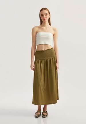 Weißes geripptes Croptop mit einem smockierten grünen Maxirock. Der Rock hat eine geraffte Textur und ein fließendes Design; getragen mit gemusterten Schuhen.