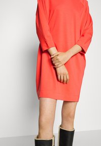 Korallenfarbenes, oversized T-Shirt-Kleid mit Dreiviertelärmeln. Weicher Stoff, minimalistisches Design, Saum endet mittig am Oberschenkel, getragen mit schwarzen Stiefeln.