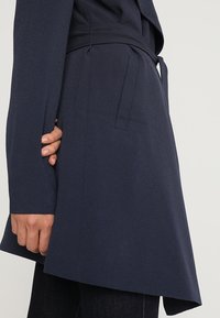 Manteau enveloppant bleu marine avec une finition texturée, doté d'une ceinture à la taille, de manches longues et d'un ourlet asymétrique.