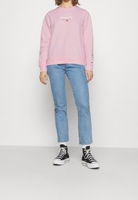 Vaaleanpunainen collegepaita, jossa on "Tommy Jeans New York" -logo, yhdistetty vaaleansinisiiin farkkuihin, joissa on repsottavat lahkeet, ja mustiin korkeavartisiin tennareihin.