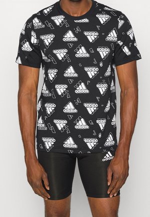 Mann trägt ein schwarzes Adidas T-Shirt mit weißen verstreuten Logos und schlichte schwarze Adidas Shorts mit Logo am linken Oberschenkel.