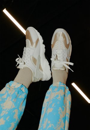 Personne portant des baskets blanches et beiges avec des chaussettes beiges et un pantalon bleu à motifs, jambes levées sur un fond sombre avec des lumières vives.