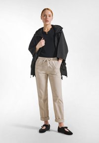 Chaqueta rompevientos negra de corte holgado, combinada con pantalones beige de cintura elástica con bolsillos delanteros. Zapatos planos negros completan el conjunto.