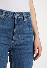 Vaqueros de mezclilla azul de cintura alta con diseño de cinco bolsillos, que cuentan con un cierre de botón metálico y acentos de costura en naranja.