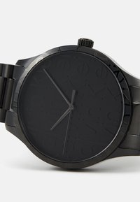 Reloj de pulsera analógico negro con acabado mate, que presenta una esfera texturizada y una correa de metal. Manecillas de hora y minuto sencillas, sin números.