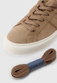 Κοντινό πλάνο καφέ σουέντ αθλητικού παπουτσιού με στρογγυλεμένη λευκή λαστιχένια σόλα και αποθέματα κορδονιών σε ίδιο χρώμα, με μπλε ετικέτα Hogan σε λευκό φόντο.