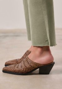 Mules à talons tissés marron avec un bout pointu et un talon noir robuste, associés à un pantalon cropped vert clair avec un ourlet effiloché.