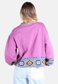 Sweatshirt violet avec un ourlet à motifs bleu, jaune et blanc. Présente des manches larges et un col rond, avec un logo dans le dos.