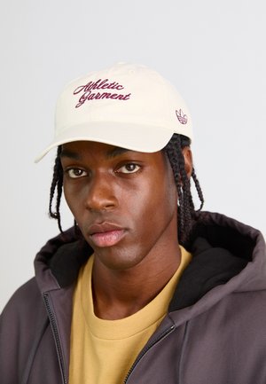 Young man with braided hair wearing a cream cap with "Athletic Garment" text, yellow shirt, and dark zip-up hoodie against a plain background.