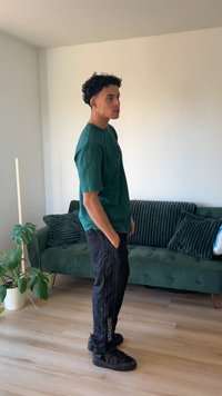 Zielona koszulka z krótkim rękawem, czarne spodnie typu cargo z bocznym logo oraz czarne trampki. Wnętrze z zieloną sofą i roślinami.