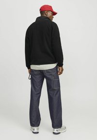 Sort fleece-trøje med høj krave, samsat med blå denimbukser med lappe-lommer og kontrasterende syninger, samt sølvfarvede sneakers.