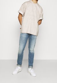 Camiseta oversize color beige claro con diseño gráfico en relieve, mangas enrolladas, combinada con jeans azul claro y zapatillas blancas.