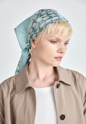 Blondynka w niebieskiej wzorzystej chustce na głowie, beżowym płaszczu i białej prążkowanej bluzce, patrząca w dół i na bok na tle białego tła.