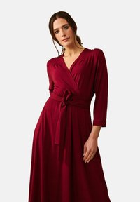 En kvinna med långt hår bär en vinröd omwrapklänning med knytmidja, poserande mot en enkel bakgrund och utstrålar självförtroende och elegans.