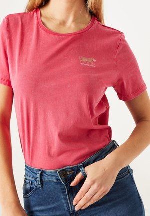 Kobieta ubrana w wyblakły czerwony T-shirt z małym logo tygrysa i napisem, w połączeniu z wysokim stanem niebieskich dżinsów, dłoń spoczywa blisko pasa.