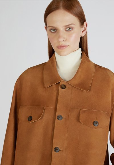 Veste en daim marron avec un col classique, fermeture à boutons sur le devant, deux poches poitrine et une texture douce. Portée par-dessus un col roulé crème clair.
