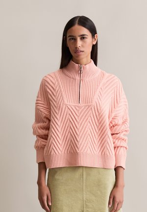 Femme portant un pullover en maille côtelée rose avec un col zippé moitié et une jupe en velours côtelé vert clair, se tenant devant un fond uni.