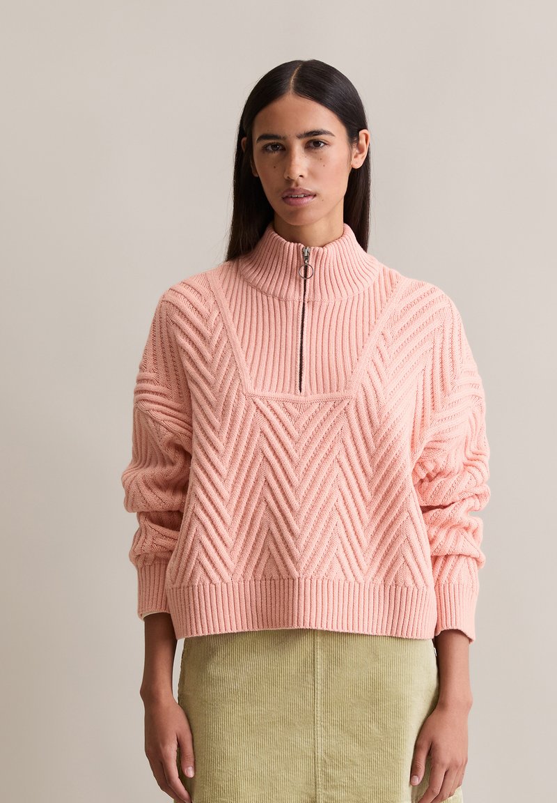 Femme portant un pullover en maille côtelée rose avec un col zippé moitié et une jupe en velours côtelé vert clair, se tenant devant un fond uni.
