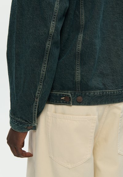 Persona con una chaqueta de mezclilla azul oscuro y pantalones beige claros con grandes bolsillos traseros, con la mano relajada a su lado.