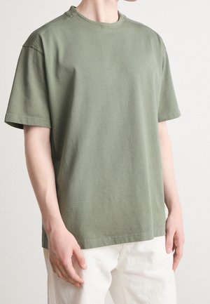 Mężczyzna ubrany w gładki oliwkowozielony T-shirt z krótkim rękawem z małym napisem „BOSS” na kołnierzyku oraz białe spodnie, stoi na jasnym tle.