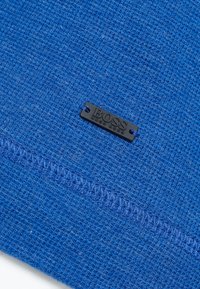 Blå texturerad tyg med ett vävt mönster, med en svart metalltagg som visar "BOSS" nära sömmen. Distinkt sömnadsdetalj synlig.