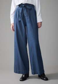 Pantalones de mezclilla de pierna ancha en azul oscuro con cintura con cinturón, que cuentan con una textura suave y dobladillo cosido, combinados con botas negras de tacón.