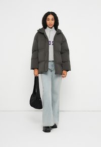 Mujer con una chaqueta acolchada oscura, suéter gris de media cremallera, jeans de pierna ancha de color azul claro, zapatos negros, sosteniendo un bolso negro de hombro.