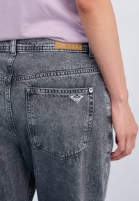 Jeans de mezclilla gris de cintura alta, con un parche de logo de cuero y un pequeño logo de corazón en el bolsillo trasero. Textura suave y corte clásico.